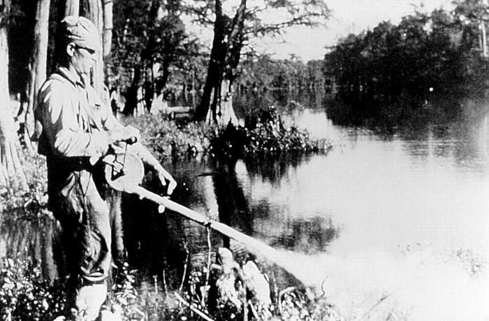 A person stands on a river bank spraying chemicals into the river to control mosquito breeding.