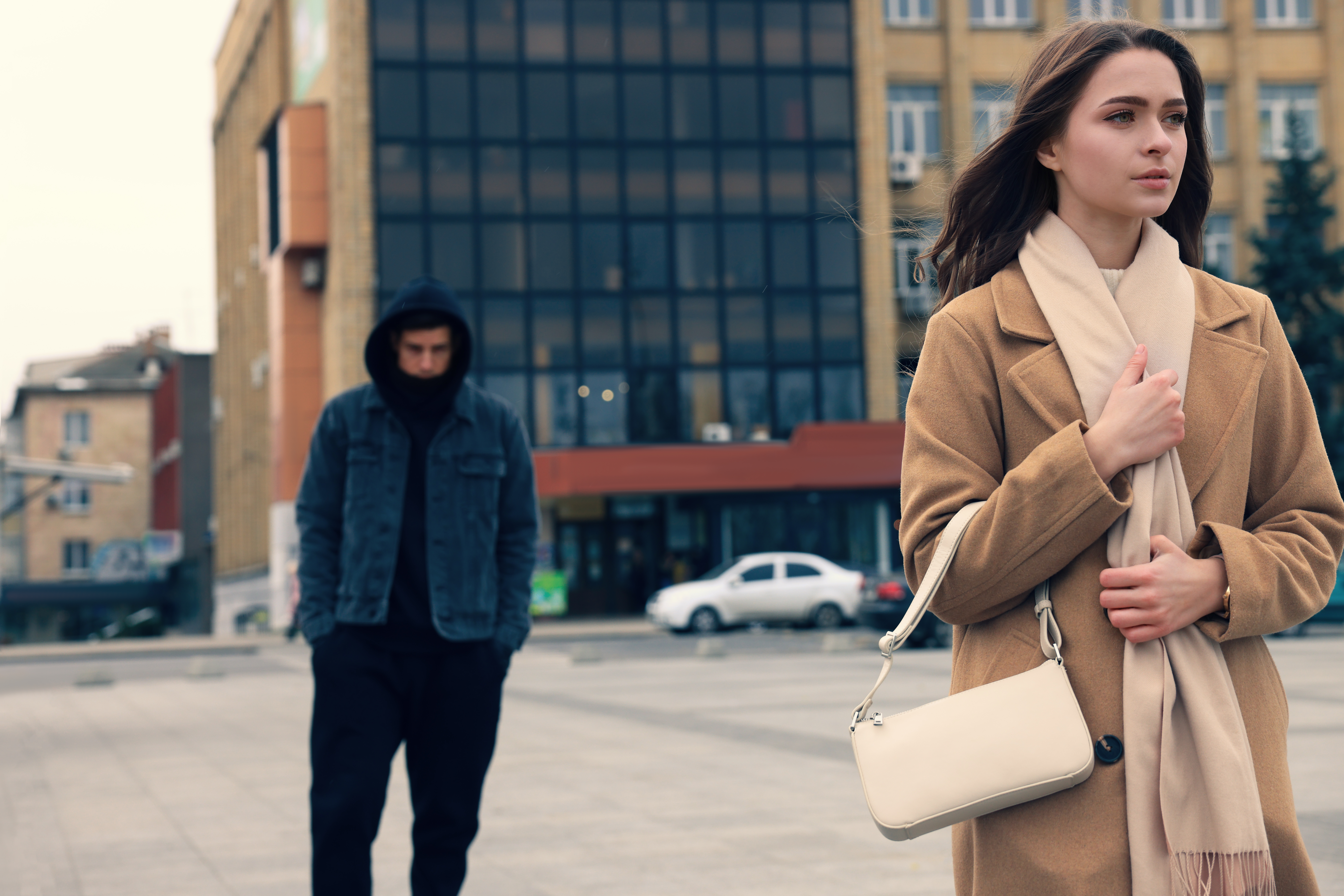 A young woman with long brown hair, wearing a tan coat and a beige scarf, walks confidently in an urban setting, holding a white handbag. In the background, a man in dark clothing and a hoodie walks behind her, appearing to follow her at a distance. The scene seems to suggest an element of suspense or concern, often associated with themes of personal safety and awareness in public spaces. The setting is in front of a modern, multi-story building with glass windows, which adds to the urban atmosphere.