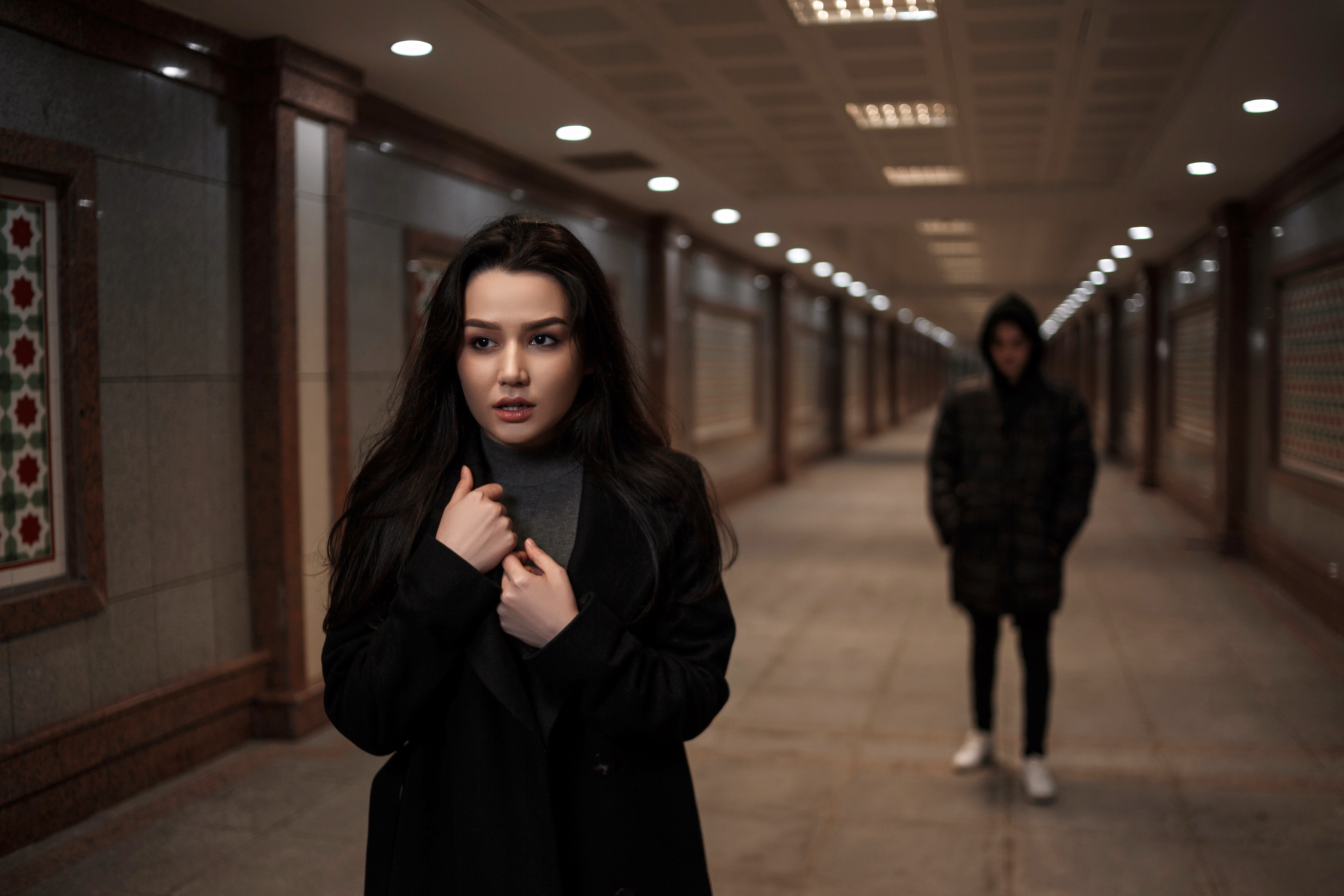 A young woman with long dark hair and wearing a black coat stands in the foreground of an empty, well-lit corridor, looking slightly anxious as she holds her coat closed. In the background, a person in a hooded jacket and white sneakers appears to be following her, adding a sense of tension and unease to the scene. The corridor's repetitive architectural details and symmetry amplify the feeling of isolation and suspense.