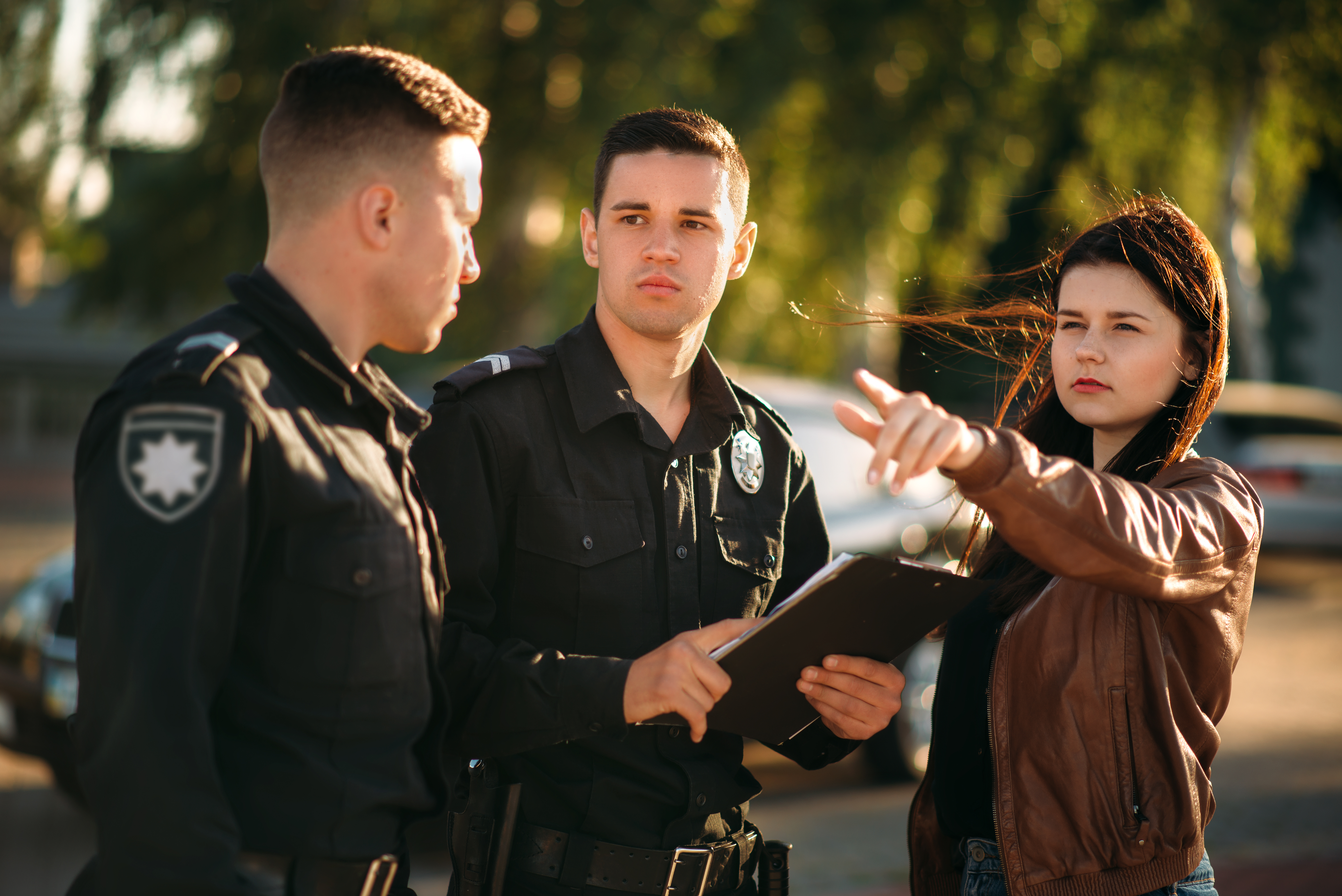 In the image, a woman is pointing to something off-camera while talking to two male police officers who are attentively listening. One officer is holding a clipboard, possibly taking notes. This scene likely depicts the woman giving a report or providing information about an incident. The background suggests they are outside, possibly at the location related to the incident.