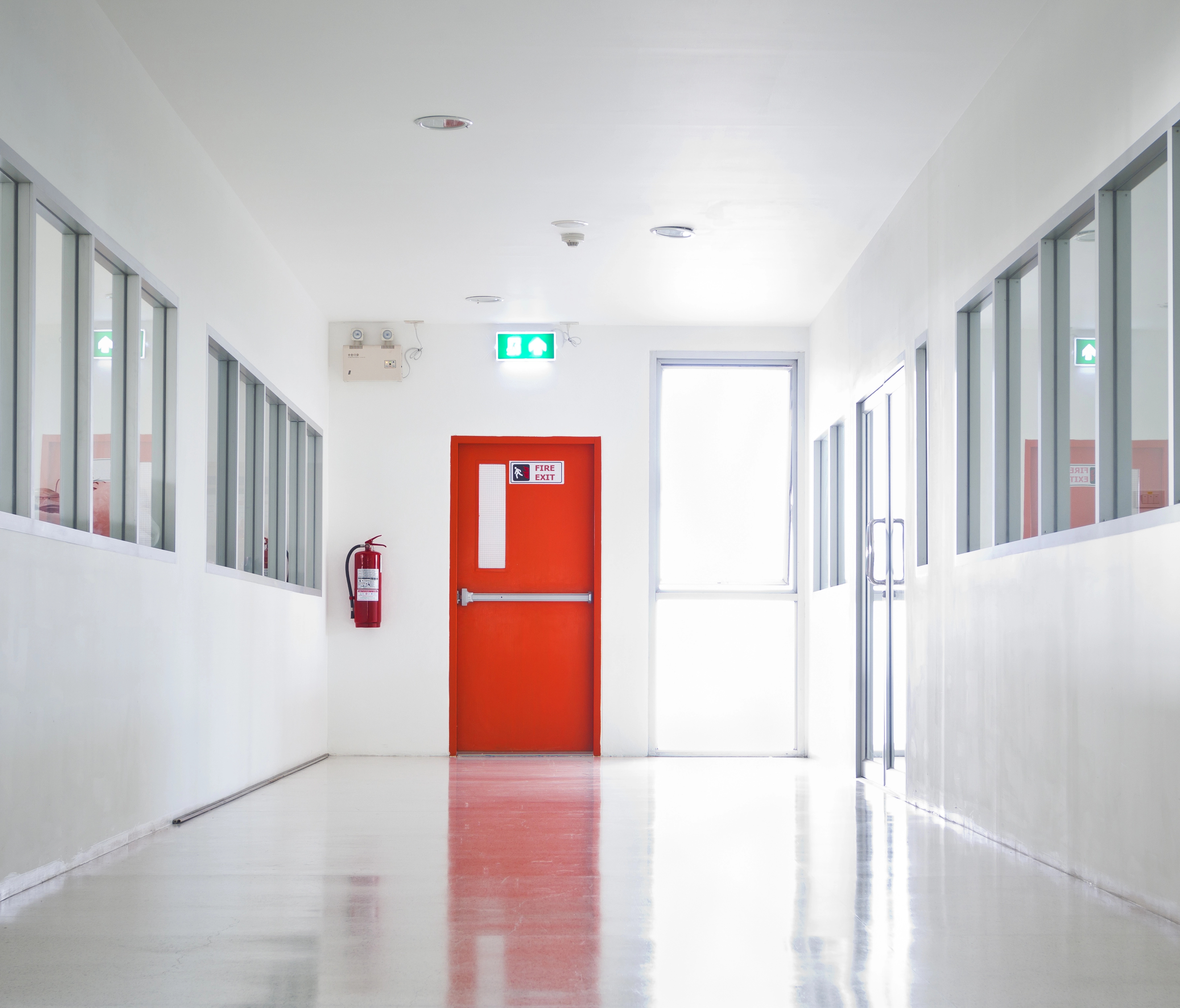 The image depicts a clean, well-lit hospital corridor. Prominently featured is a bright red emergency exit door at the end of the hallway, equipped with a "Push Bar to Open" mechanism. The corridor has several windows on both sides, allowing visibility into adjacent rooms. An emergency exit sign and a fire extinguisher are also visible next to the door, indicating adherence to safety protocols.