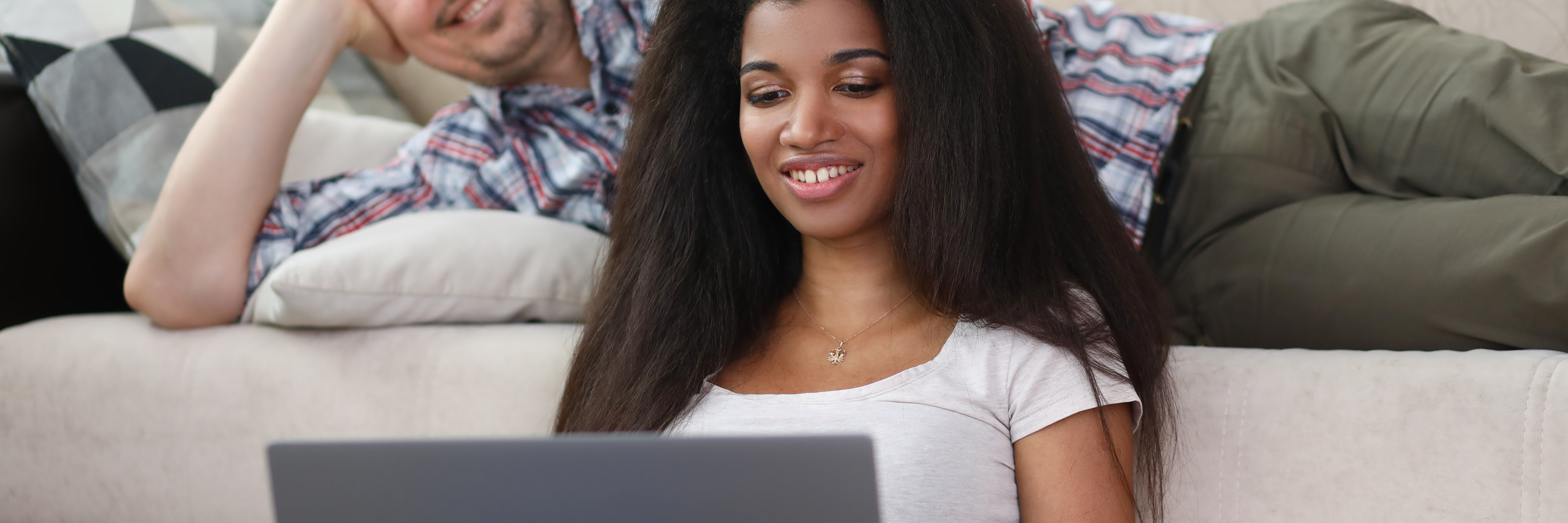 A woman with long, dark hair sits comfortably on a light-colored couch, smiling as she looks at her laptop. She is wearing a casual white t-shirt. Behind her, a man in a plaid shirt reclines on the couch, also looking relaxed. Both individuals appear content and engaged, suggesting they are enjoying a leisurely activity together.