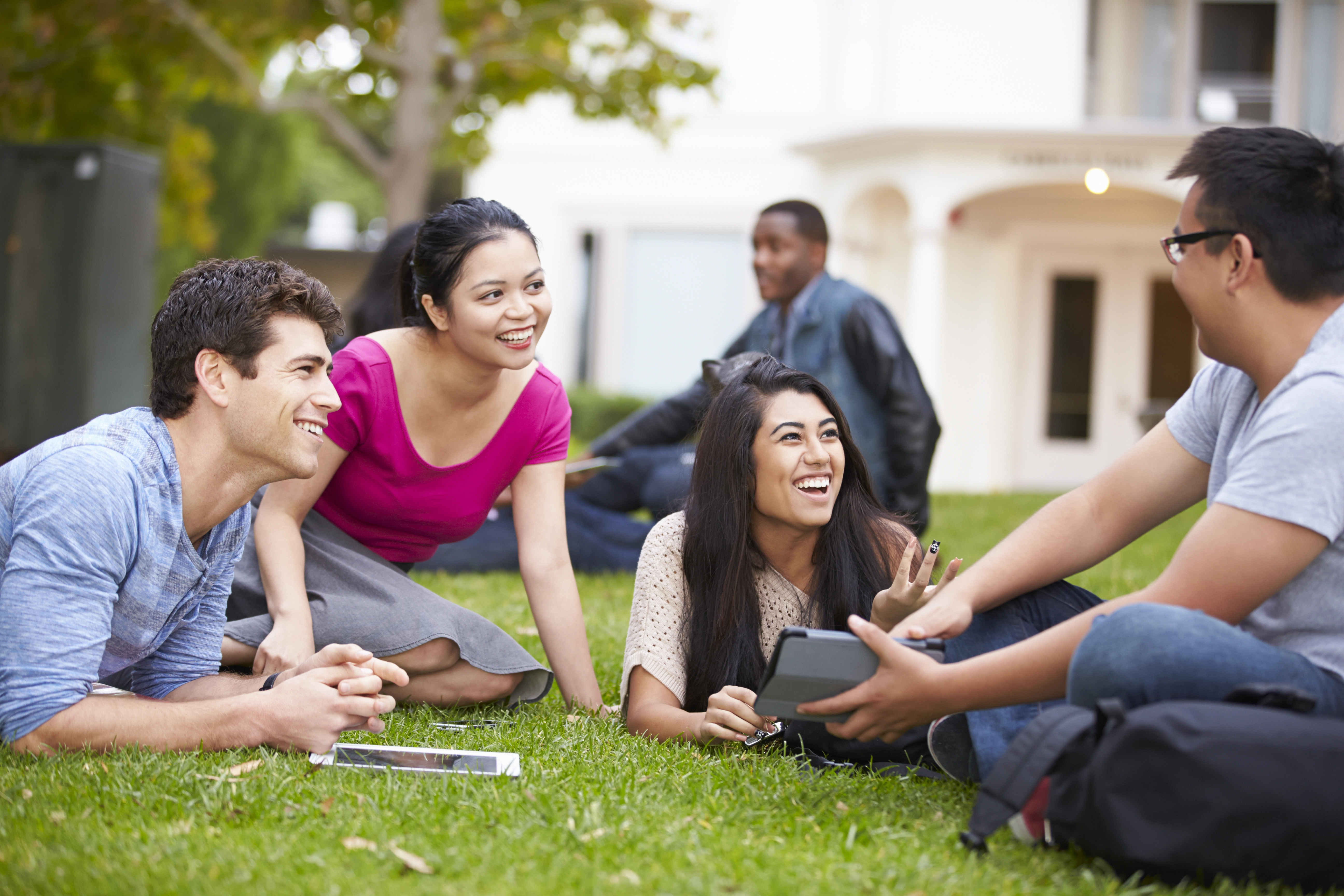 A diverse group of four college-aged students, consisting of two males and two females, are sitting and lying on a grassy lawn, engaging in an animated conversation. They appear relaxed and happy, with one male holding a tablet while the others listen and interact. The background shows a white campus building and trees, suggesting a casual, outdoor study or social session. One more student is seen sitting alone in the distance. This image illustrates the social and collaborative aspect of student life on campus.