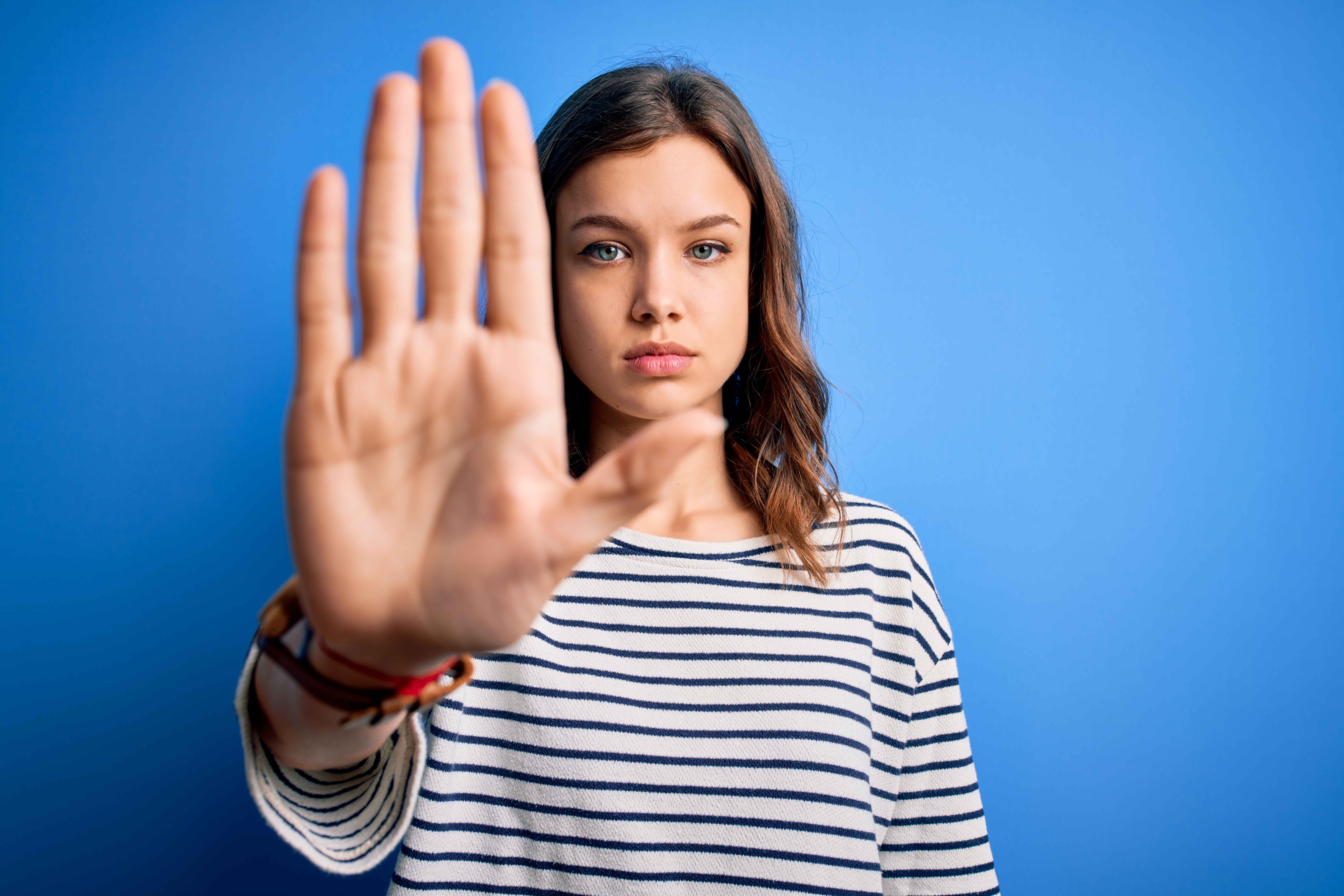 A young woman with blue eyes and brown hair, dressed in a white and black striped long-sleeve shirt, stands against a solid blue background. She faces forward with a serious expression, holding her right hand up with her palm facing outward in a "stop" gesture. This image conveys a clear message of refusal or a request to halt an action, emphasizing boundaries or opposition.