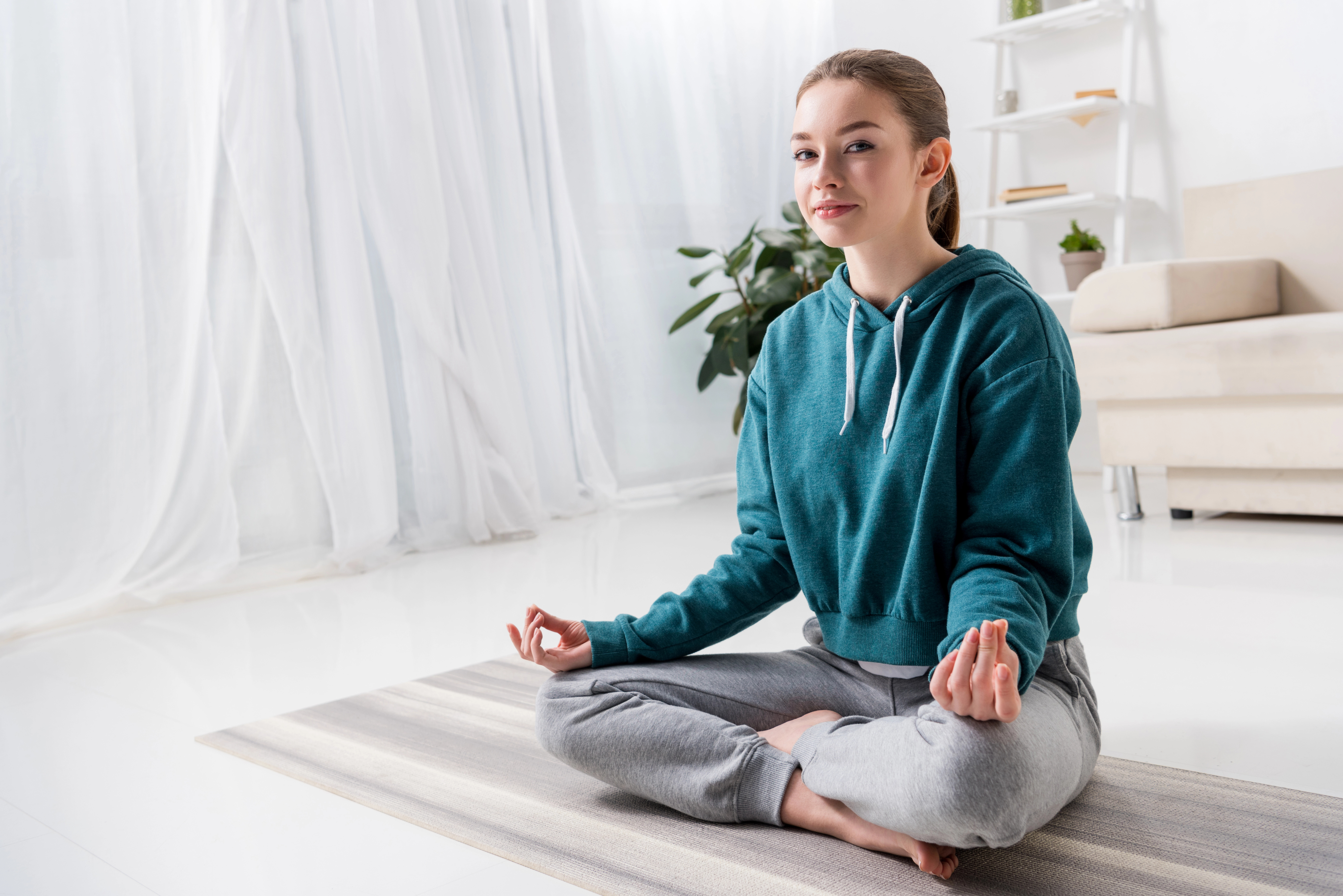 The image shows a young woman sitting cross-legged on a yoga mat in a bright, airy room. She is wearing a teal sweatshirt and grey sweatpants, and she has a relaxed, focused expression on her face. Her hands rest on her knees with palms up in a meditative position. The background includes a light-filled window with sheer curtains, a plant, a beige couch, and a shelf, all suggesting a serene home environment suitable for meditation or yoga practice.