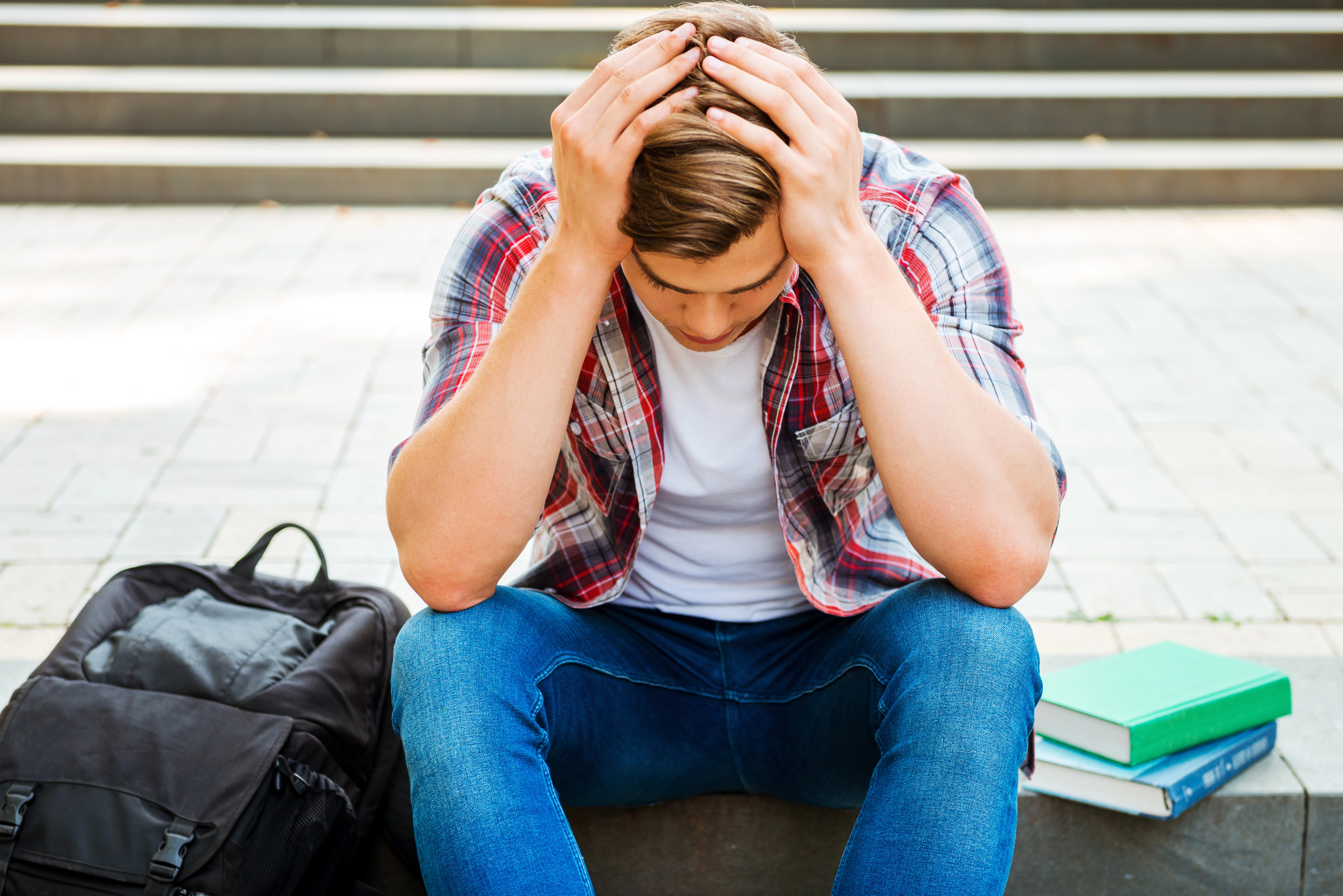 A young man is sitting with his head in his hands, looking distressed. He is wearing a plaid shirt over a white t-shirt and blue jeans. Beside him are a black backpack and two books, hinting that he might be a student dealing with academic stress or personal issues. The setting appears to be outside, near some steps, suggesting he could be on a school or college campus.