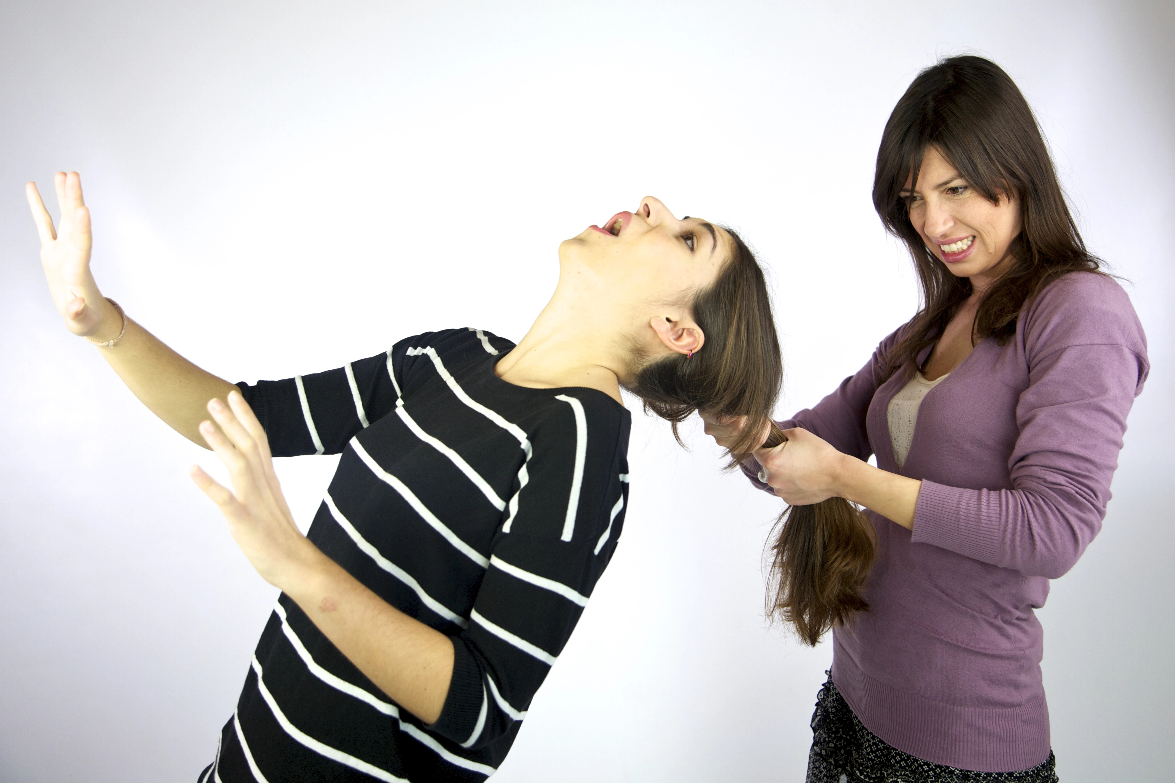 The image shows a moment of playful interaction between two women. The woman on the left, wearing a black and white striped shirt, leans back and appears to be laughing or reacting dramatically with her hands up. The woman on the right, in a lavender sweater, gently pulls on the other woman’s long hair with a mischievous smile. Their expressions and body language suggest a light-hearted and humorous scenario.